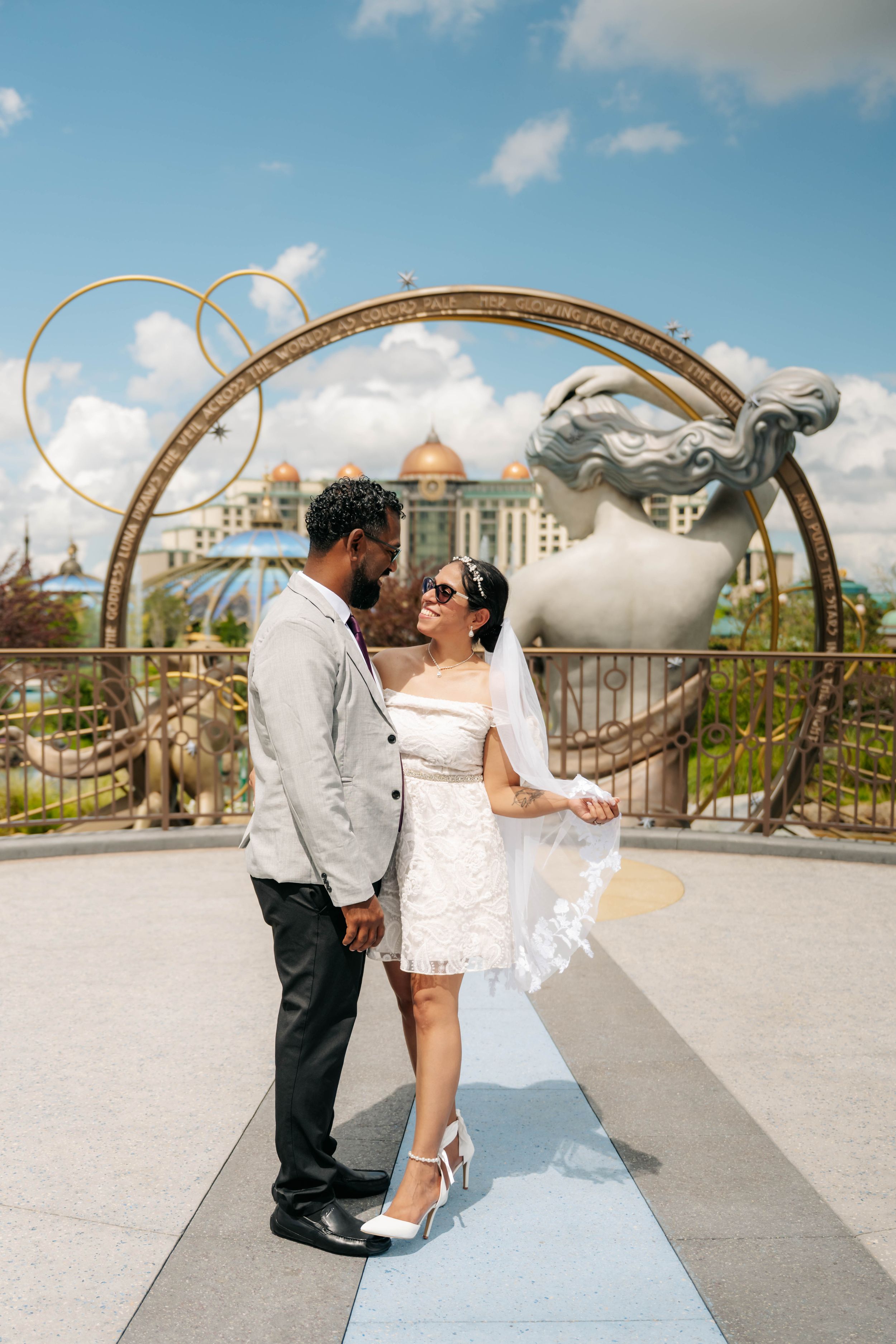 Interracial couple in wedding attire posing before ornate golden arch sculpture outdoors on sunny day. Epic universe universal orlando