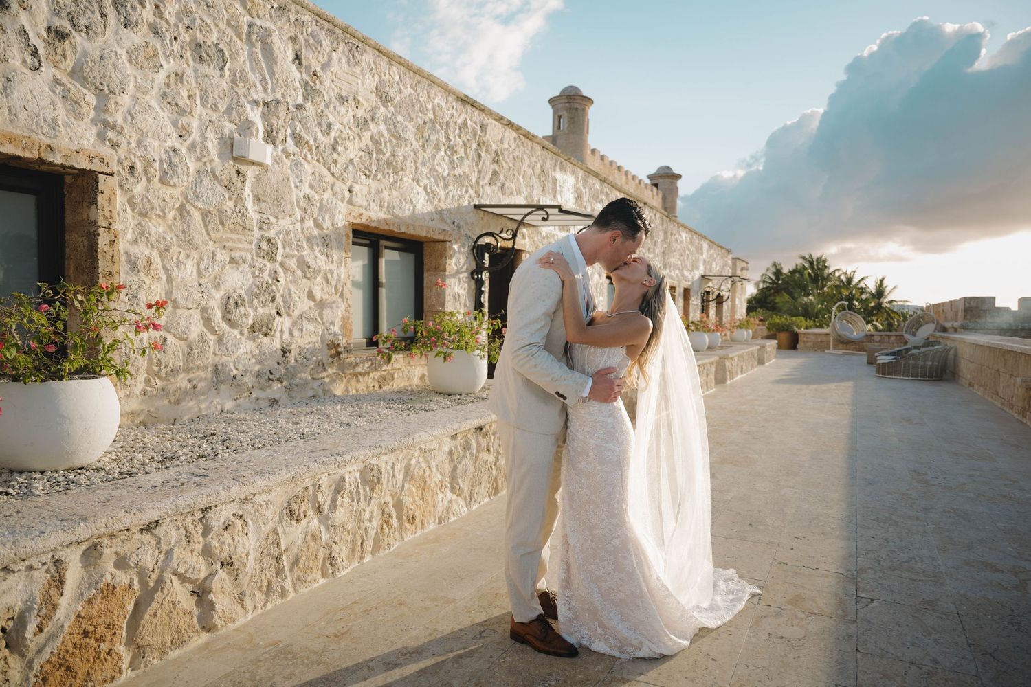 Bride and groom share a romantic kiss on a rustic stone terrace at sunset destination wedding.