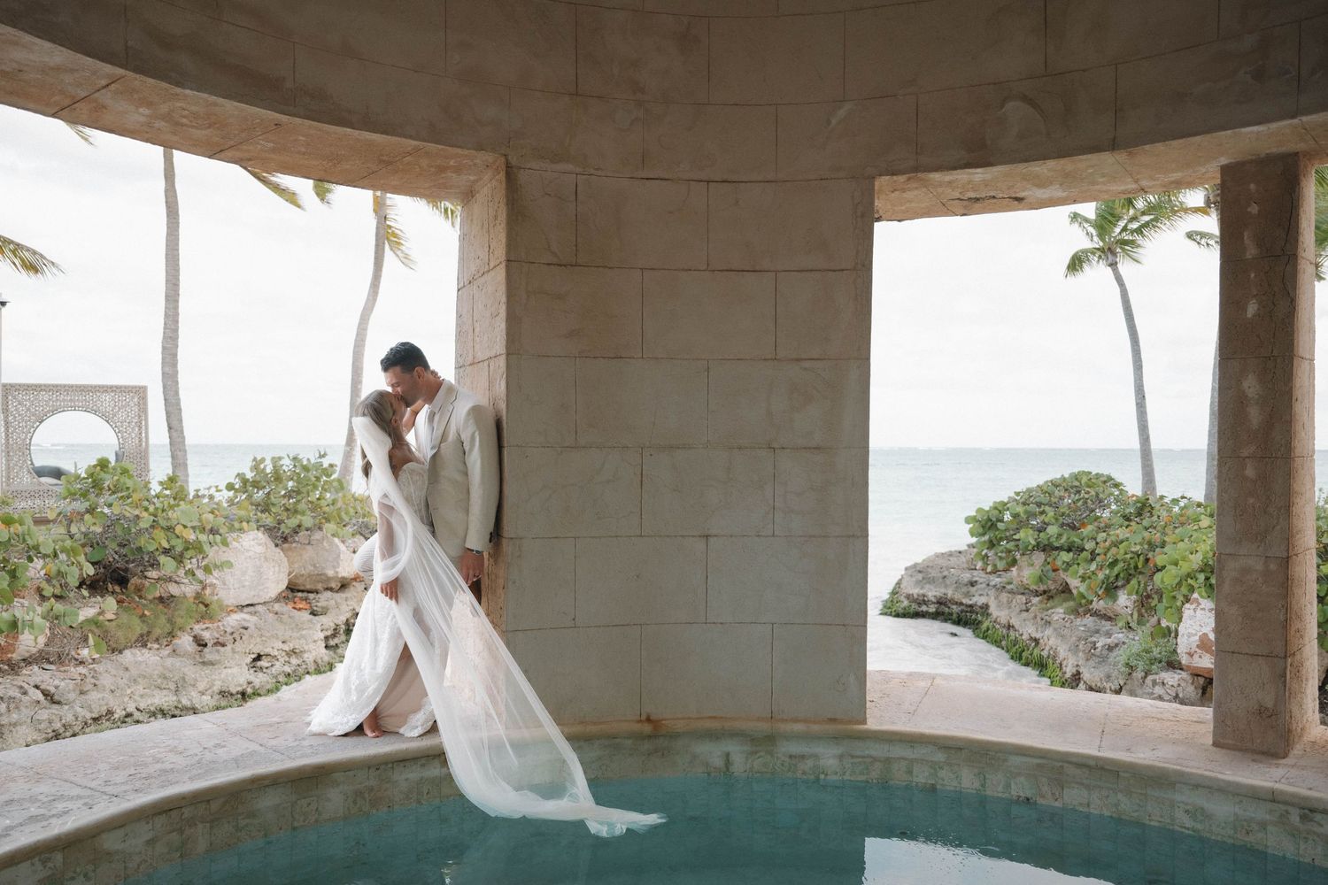 Bride and groom share a kiss beside a pool with ocean views at a luxury destination wedding.