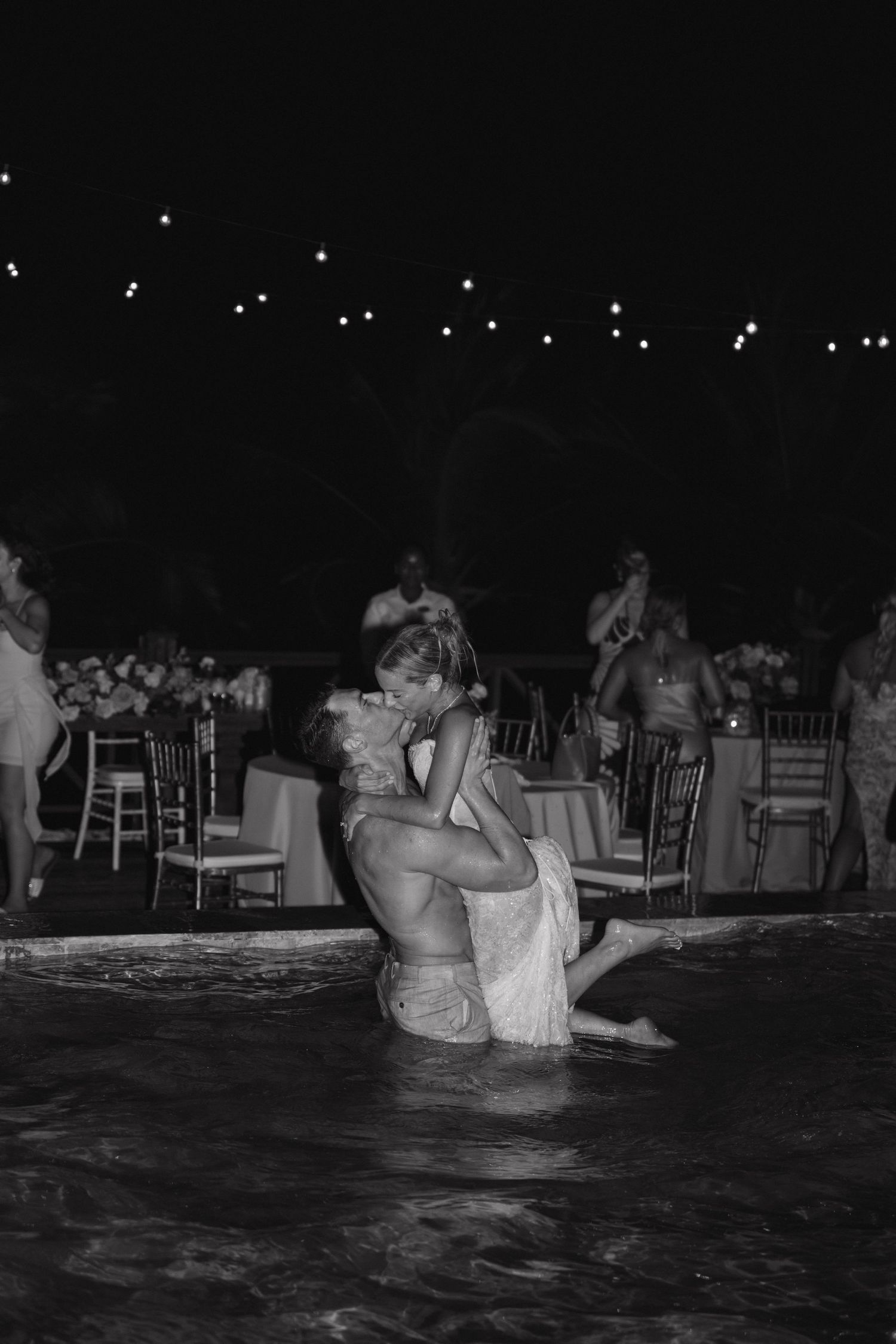 Groom lifts bride for a romantic kiss in a pool during their nighttime wedding reception.