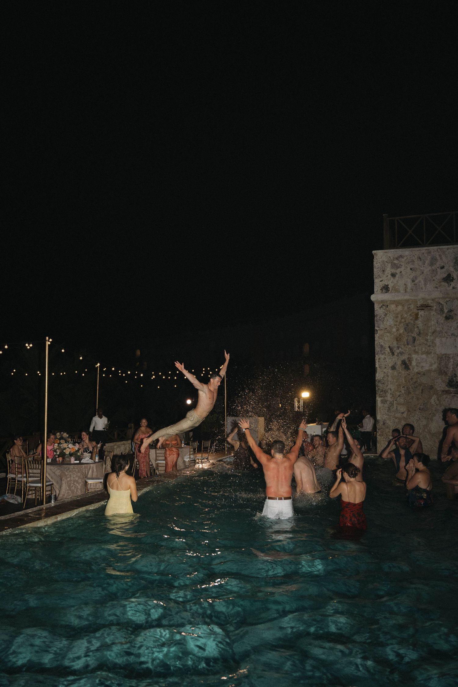Wedding guests celebrate in a pool at night with string lights glowing at a destination wedding.