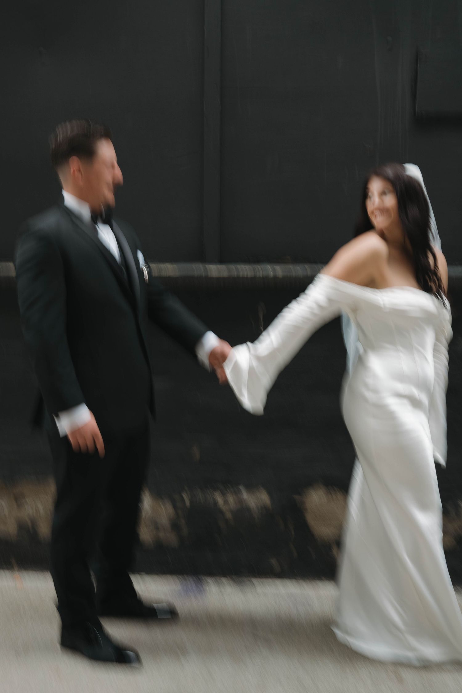 Motion-blur photo of smiling bride in off-shoulder gown and groom holding hands against a dark wall.