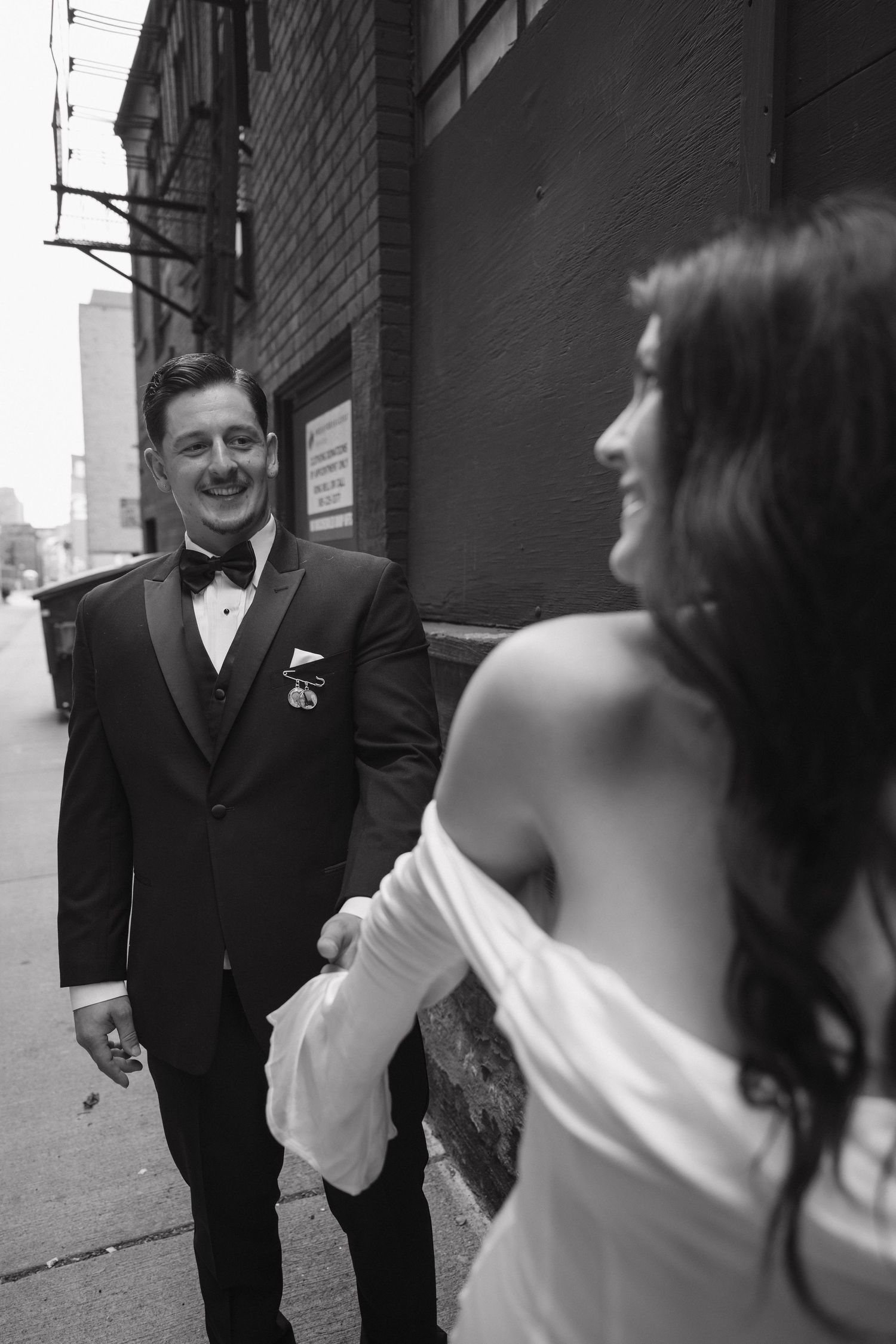 Black and white photo of smiling groom in tuxedo looking at bride in off-shoulder gown on urban street.