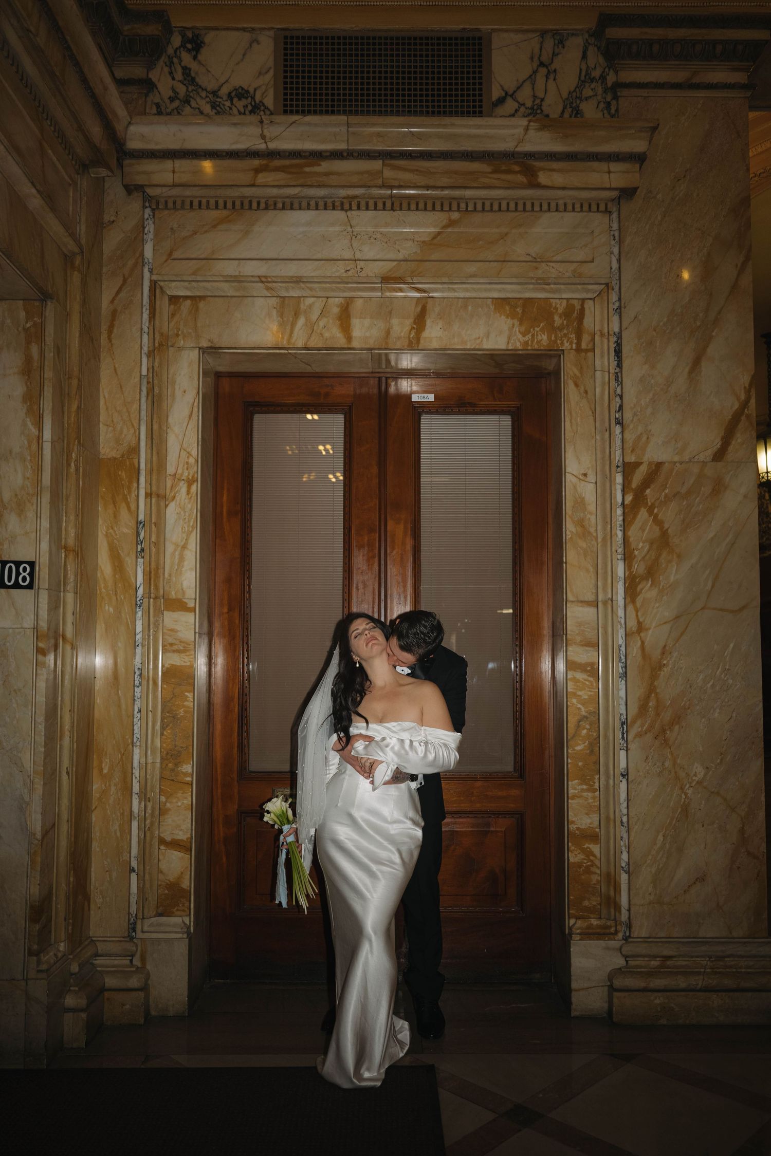 Bride in off-shoulder gown and groom share a joyful embrace before ornate marble doors at night.