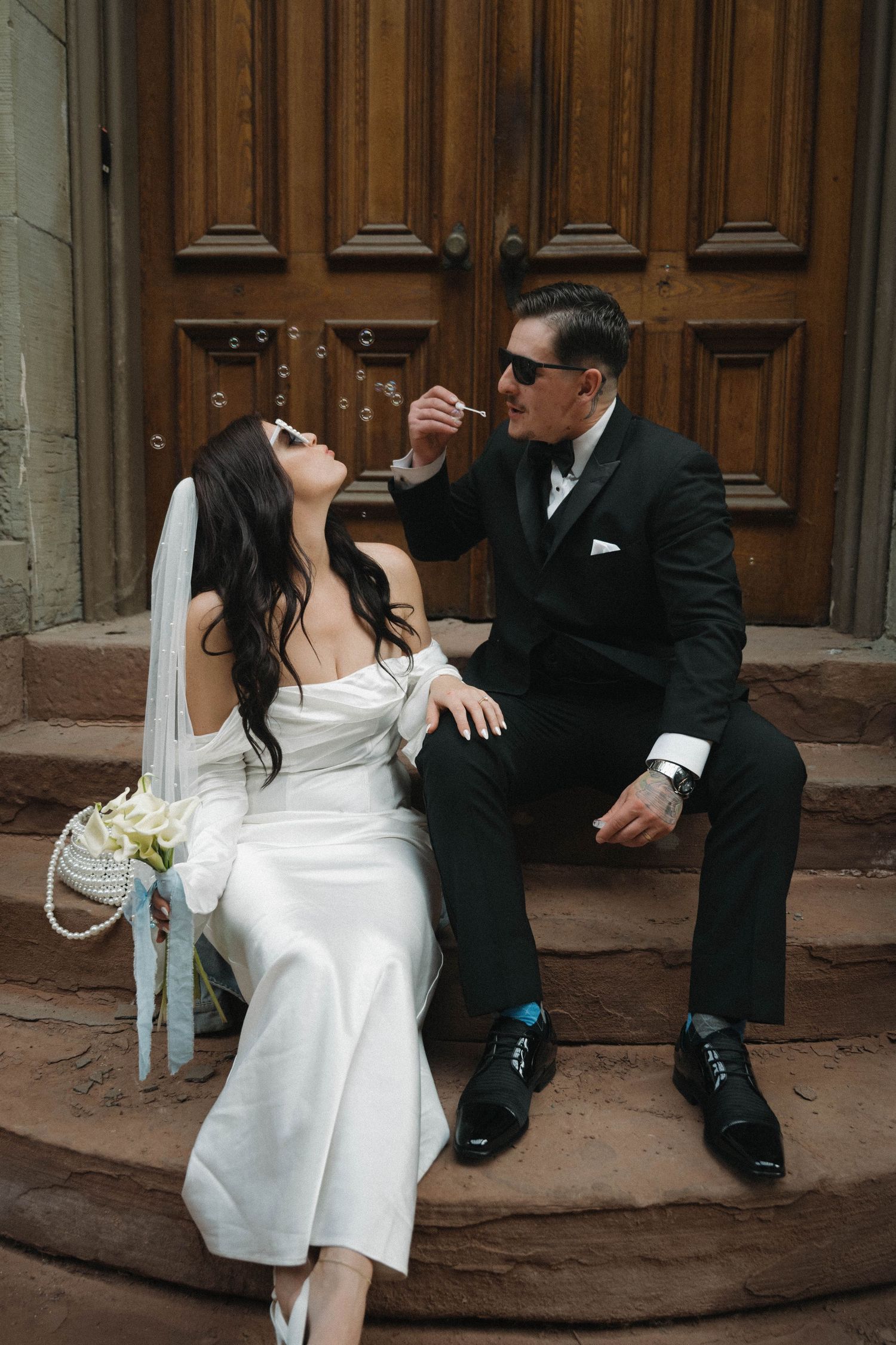 Bride and groom in sunglasses sit on church steps, groom blowing bubbles on their wedding day.