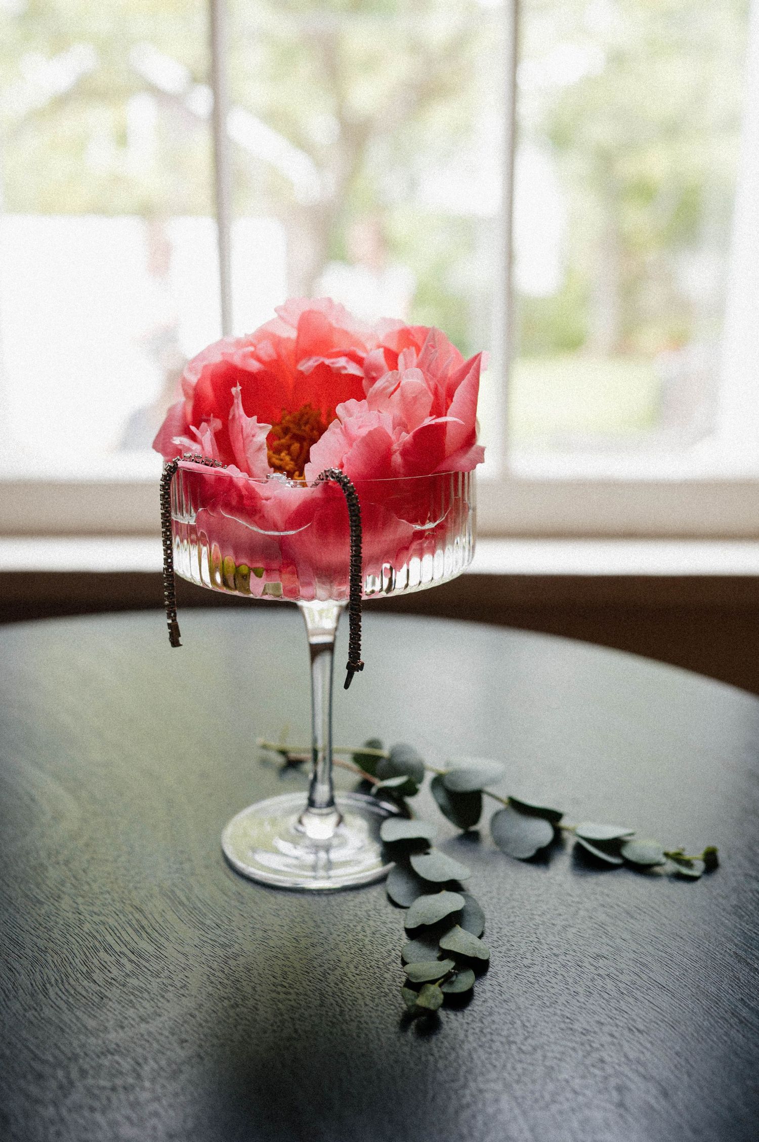 Pink peony bloom displayed in a crystal coupe glass with a delicate bracelet on a dark table.