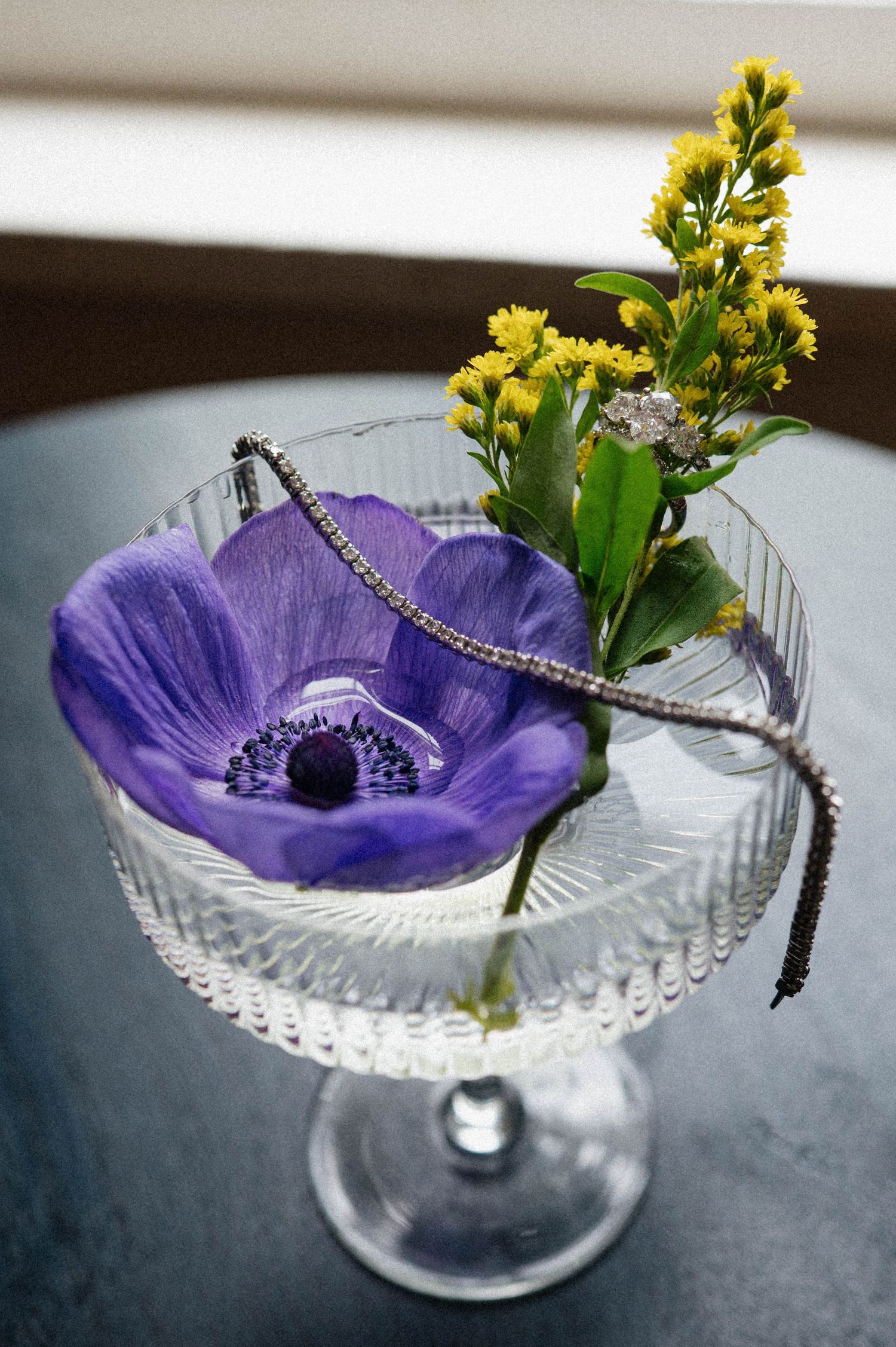 Purple anemone and yellow wildflowers arranged in a crystal coupe glass with a diamond bracelet.