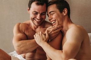 Two shirtless people laugh and embrace intimately while sitting together in a candid moment. Male boudoir photography in studio
