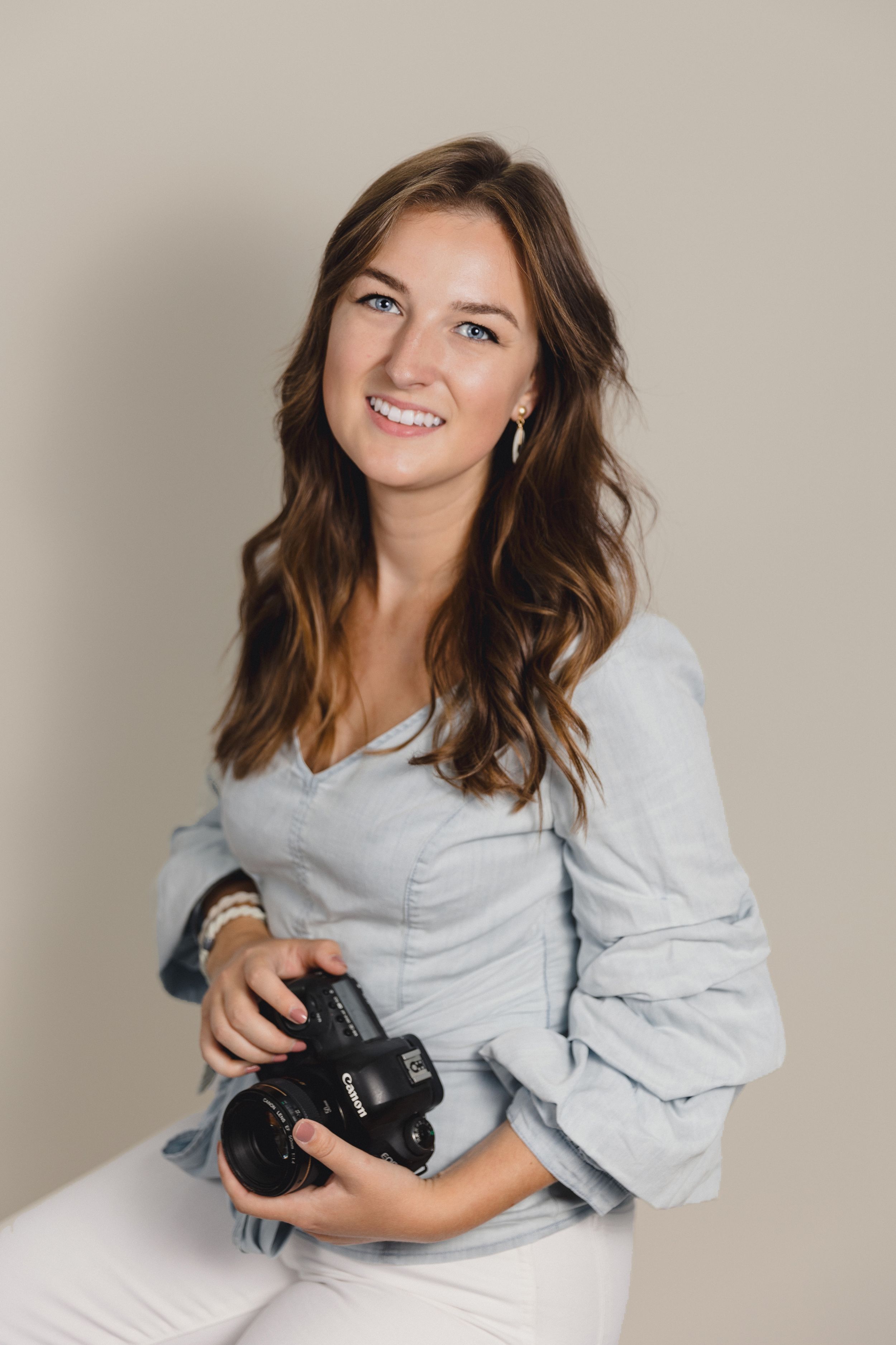 Professional photographer in light gray sweater poses with camera against neutral backdrop in studio setting.