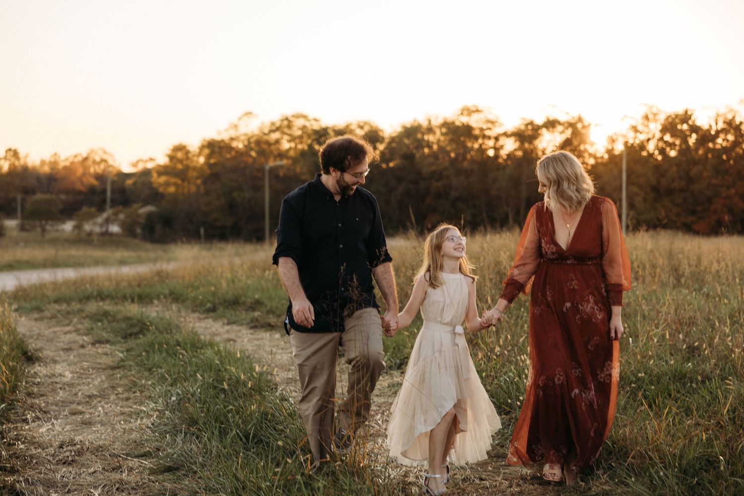 Family of three walking hand-in-hand through golden field at sunset, parents in boho attire with young girl in white dress.