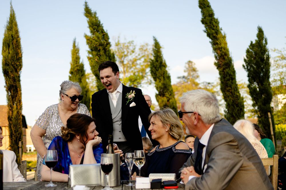 Wedding guests enjoying conversation and drinks at an outdoor table with tall cypress trees in the background.