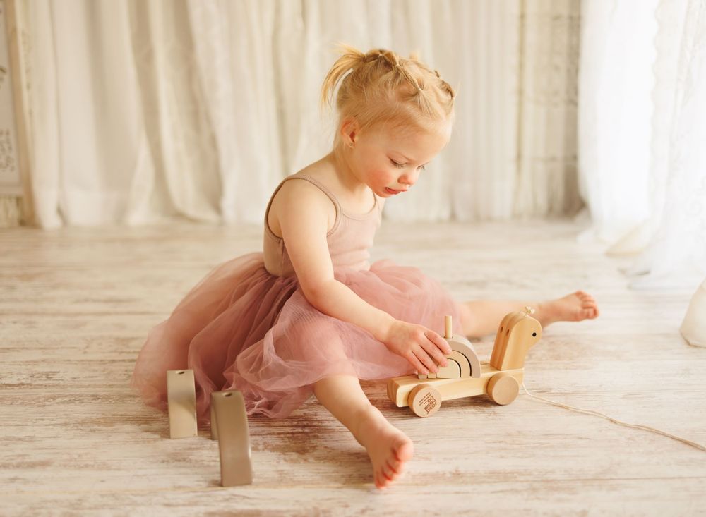 A small child in a pink tutu dress plays with a wooden toy on the floor.