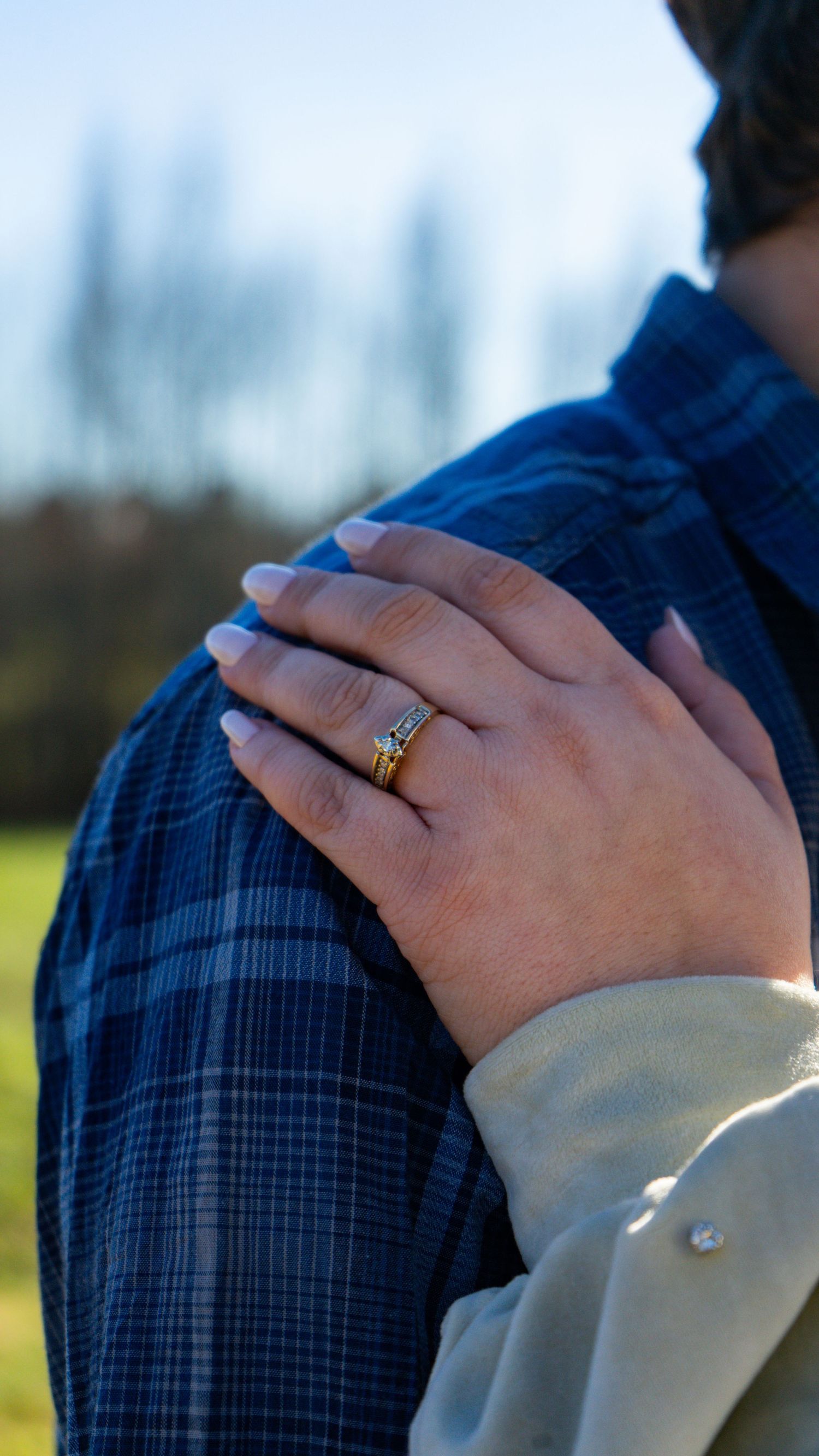 A close-up of hands showing off a wedding ring while resting on a blue plaid shirt in outdoor lighting.