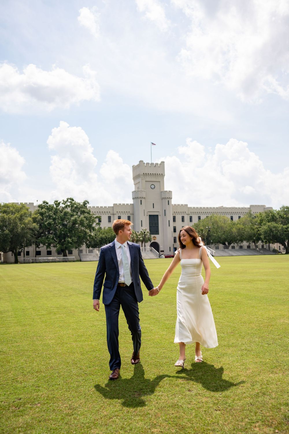 Couple in wedding attire walking and holding hands on a sunny lawn at The Citadel with white castle buildings in background.