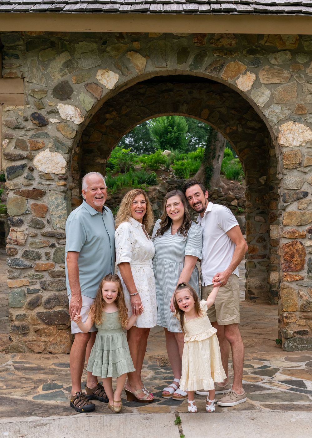 Multi-generational family of six poses under stone arch outdoors, smiling together in coordinated pastel outfits.