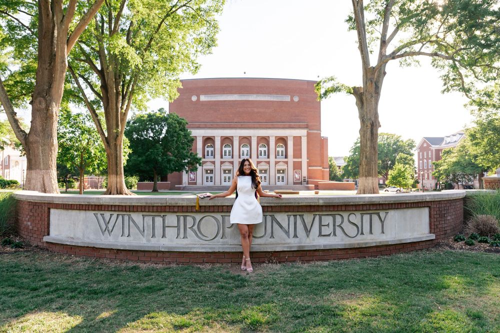 Woman in white dress posing in front of Winthrop University sign with brick building and trees in background.