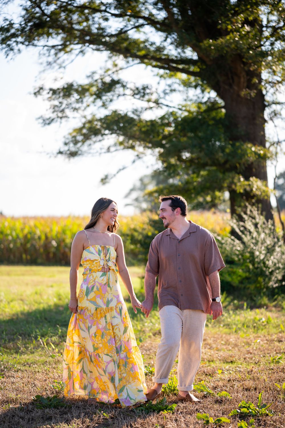 A couple walks hand in hand through a sunlit field wearing summer attire near a cornfield.