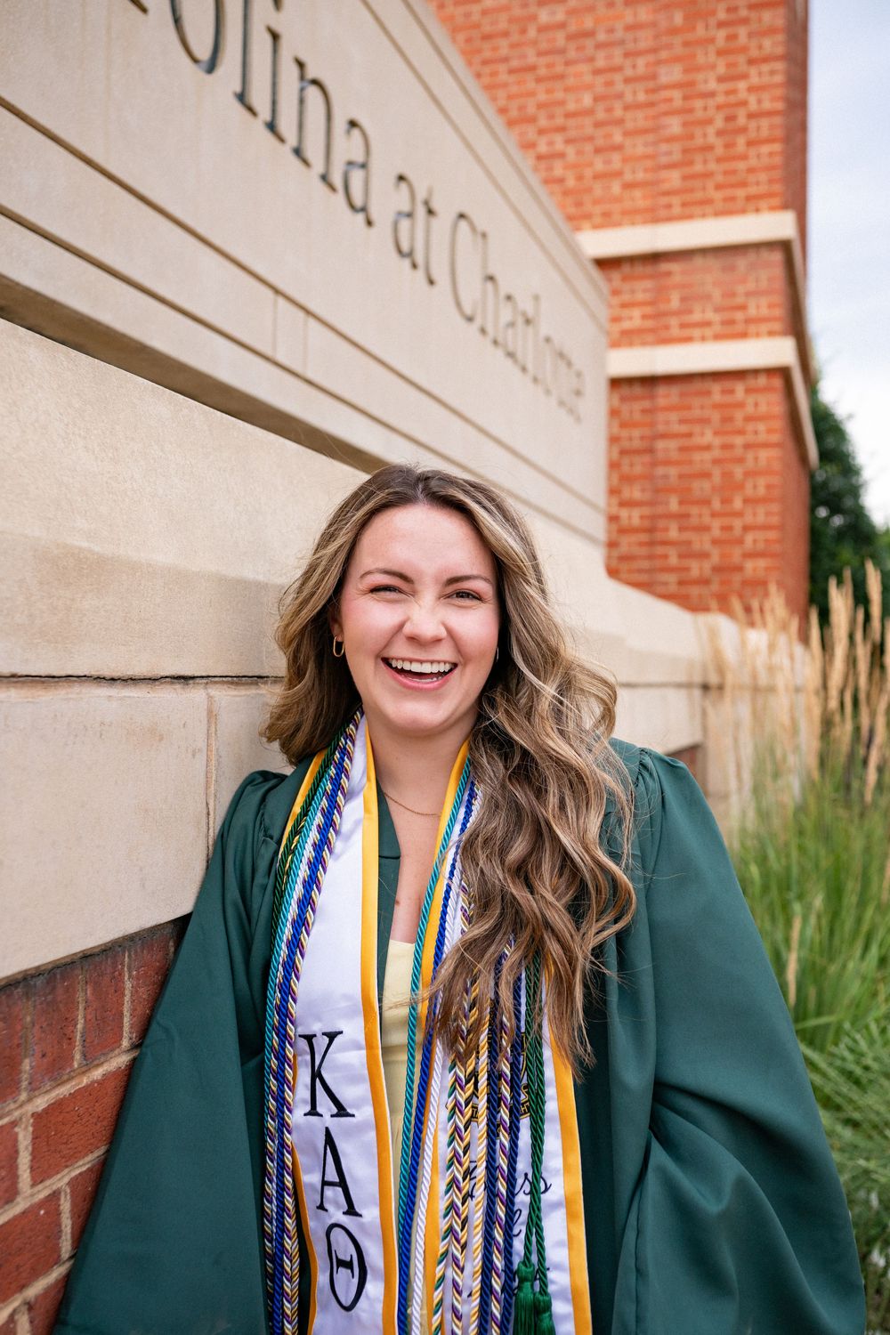 Graduate wearing green commencement gown and decorated stoles poses smiling against brick wall during graduation ceremony.