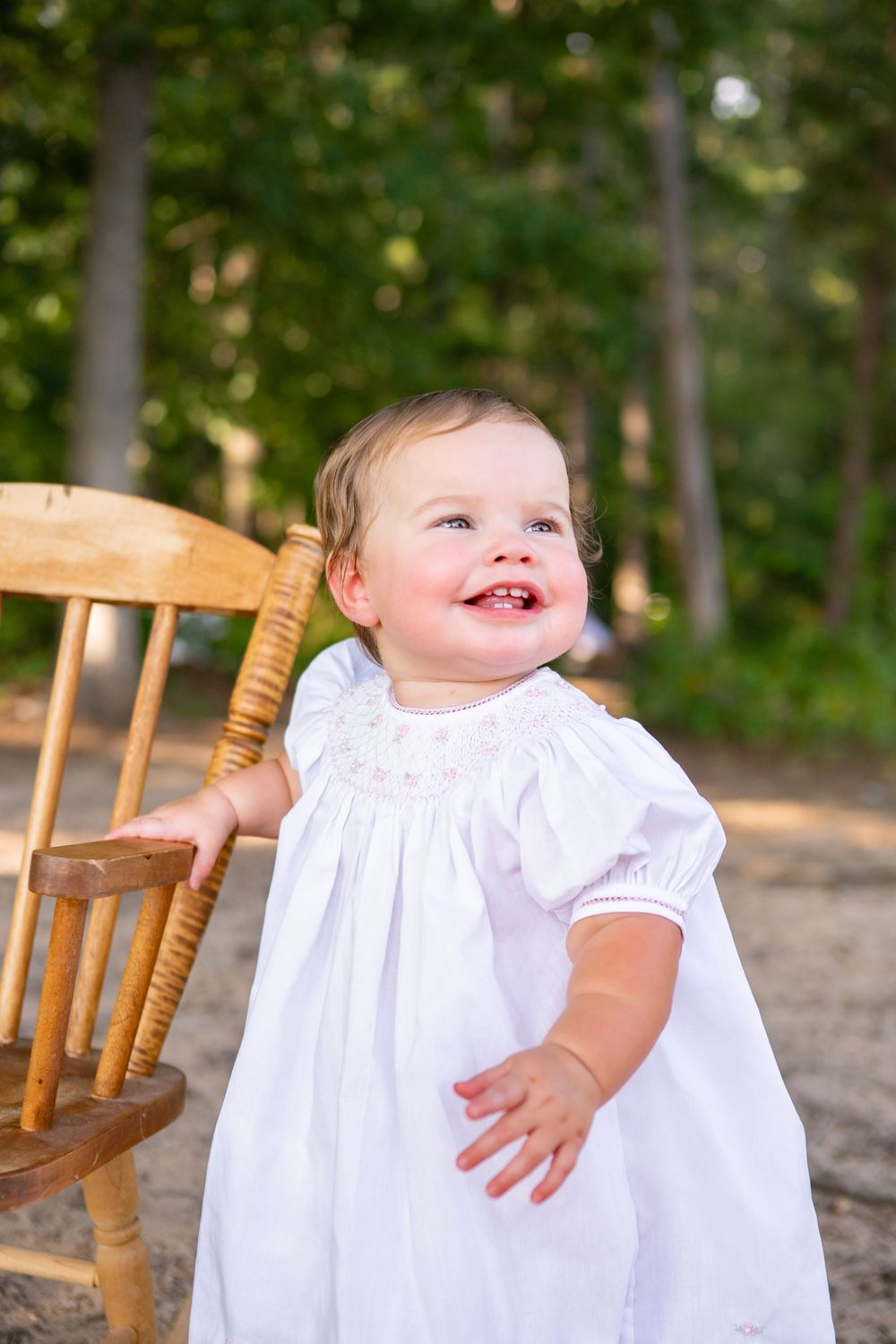 A baby in a white dress sits on a vintage wooden rocking chair against a leafy green outdoor background.