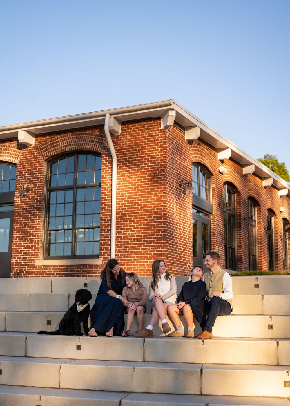 A group sits together on concrete steps outside a brick building with arched windows during golden hour lighting.