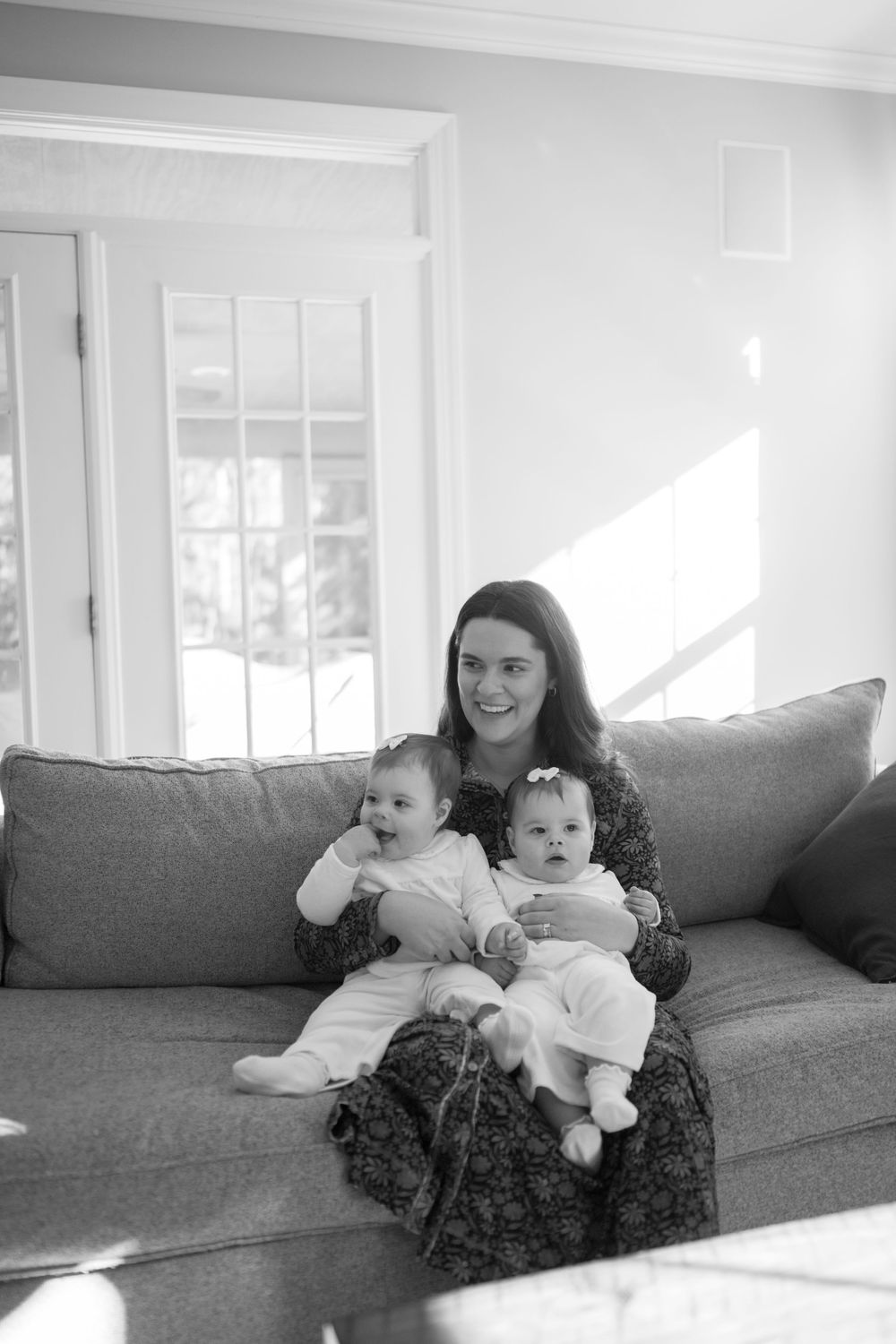 A series of black and white photos showing a parent and baby relaxing together on a gray couch in sunlit room.
