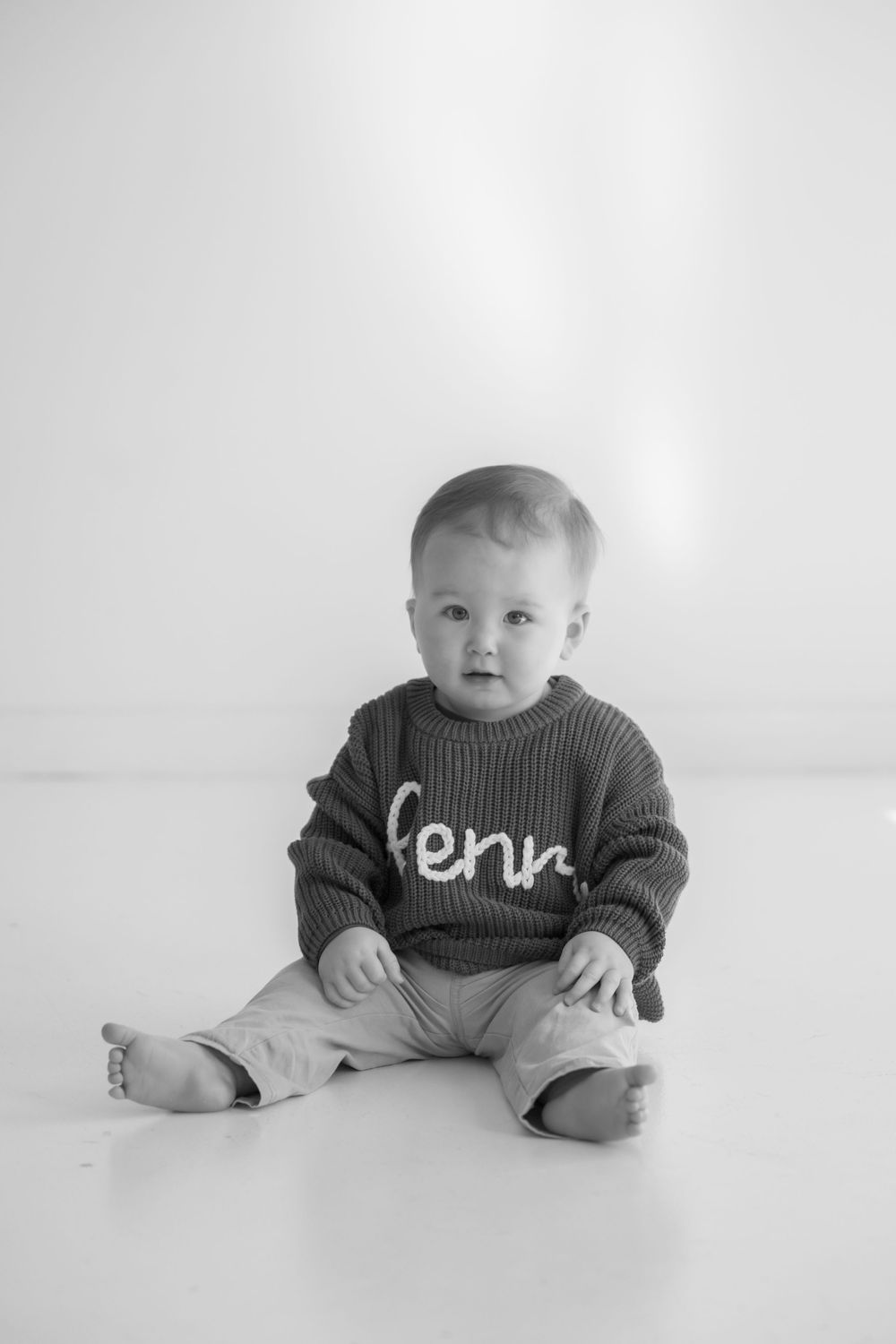 Four black and white photos of a baby sitting on a white backdrop wearing a knit sweater, shown in different poses.