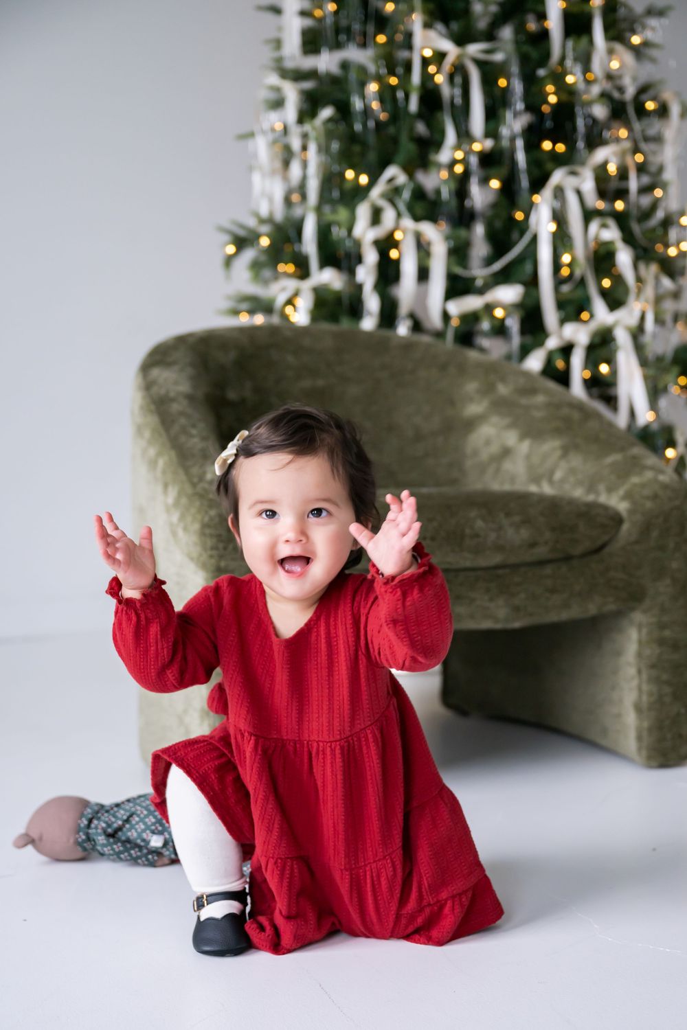 A baby in a red holiday dress sits on the floor in front of a decorated Christmas tree with twinkling lights.