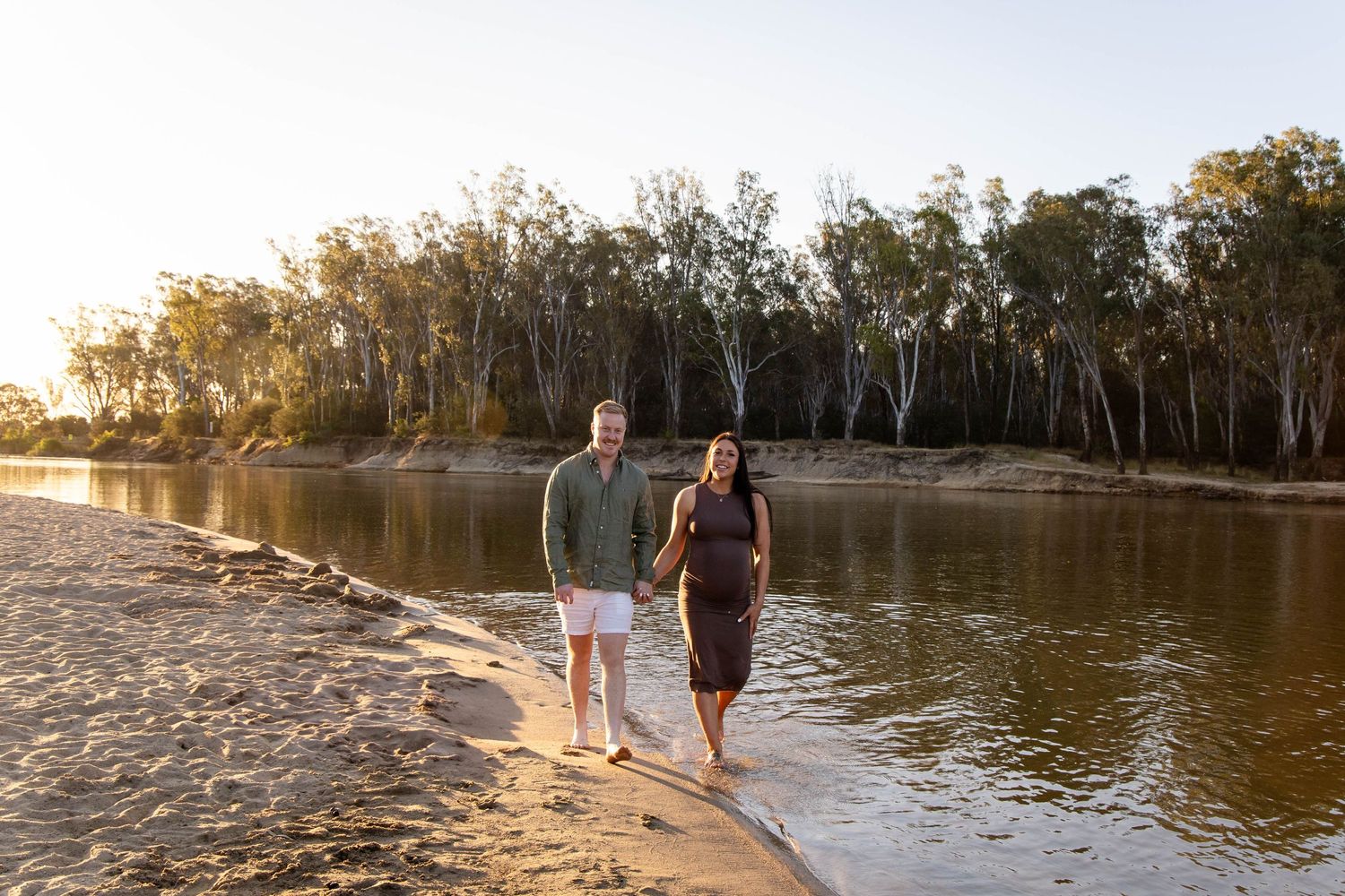 Two people walking along a sandy riverbank at sunset with eucalyptus trees lining the water's edge.