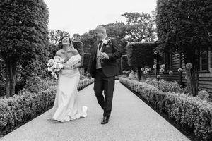 A couple in formal wedding attire walk down a garden path lined with manicured hedges.