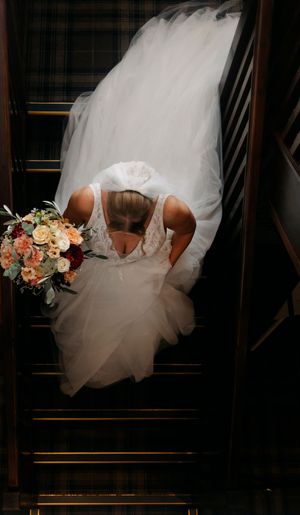 Aerial view of a bride in a flowing white wedding dress descending dark wooden stairs while holding a peach bouquet.