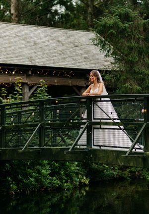A bride in a white dress stands on a rustic wooden bridge surrounded by lush greenery and string lights.