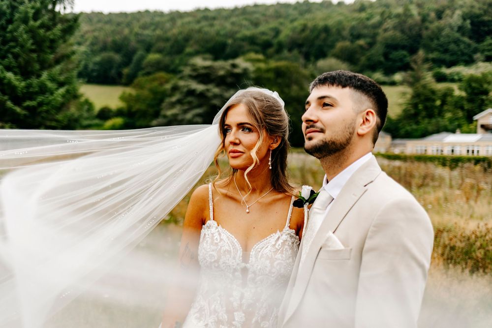 A couple poses in formal wedding attire outdoors as the bride's veil flows dramatically in the wind.