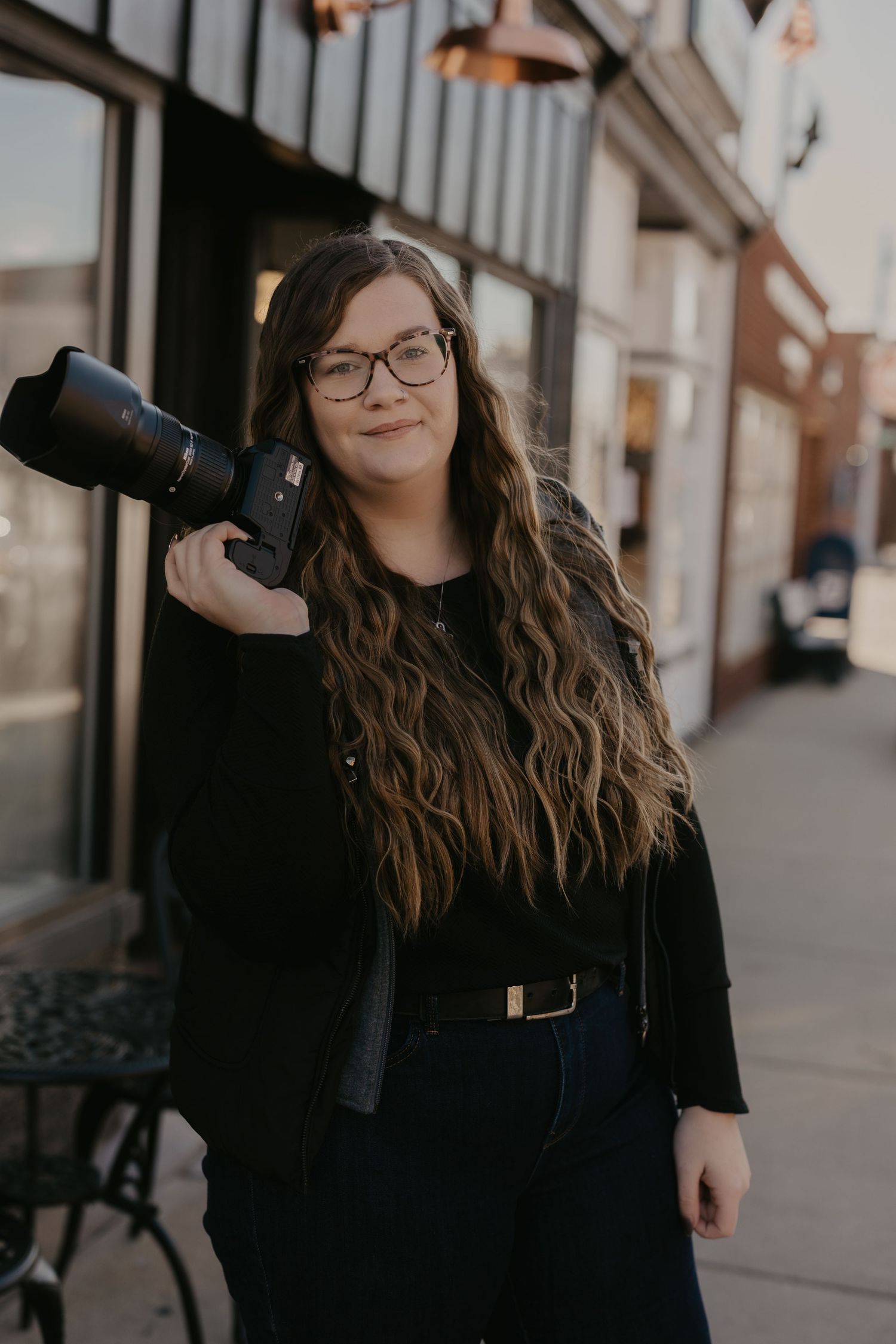 Female photographer with long wavy hair and glasses holds camera on city sidewalk at dusk.