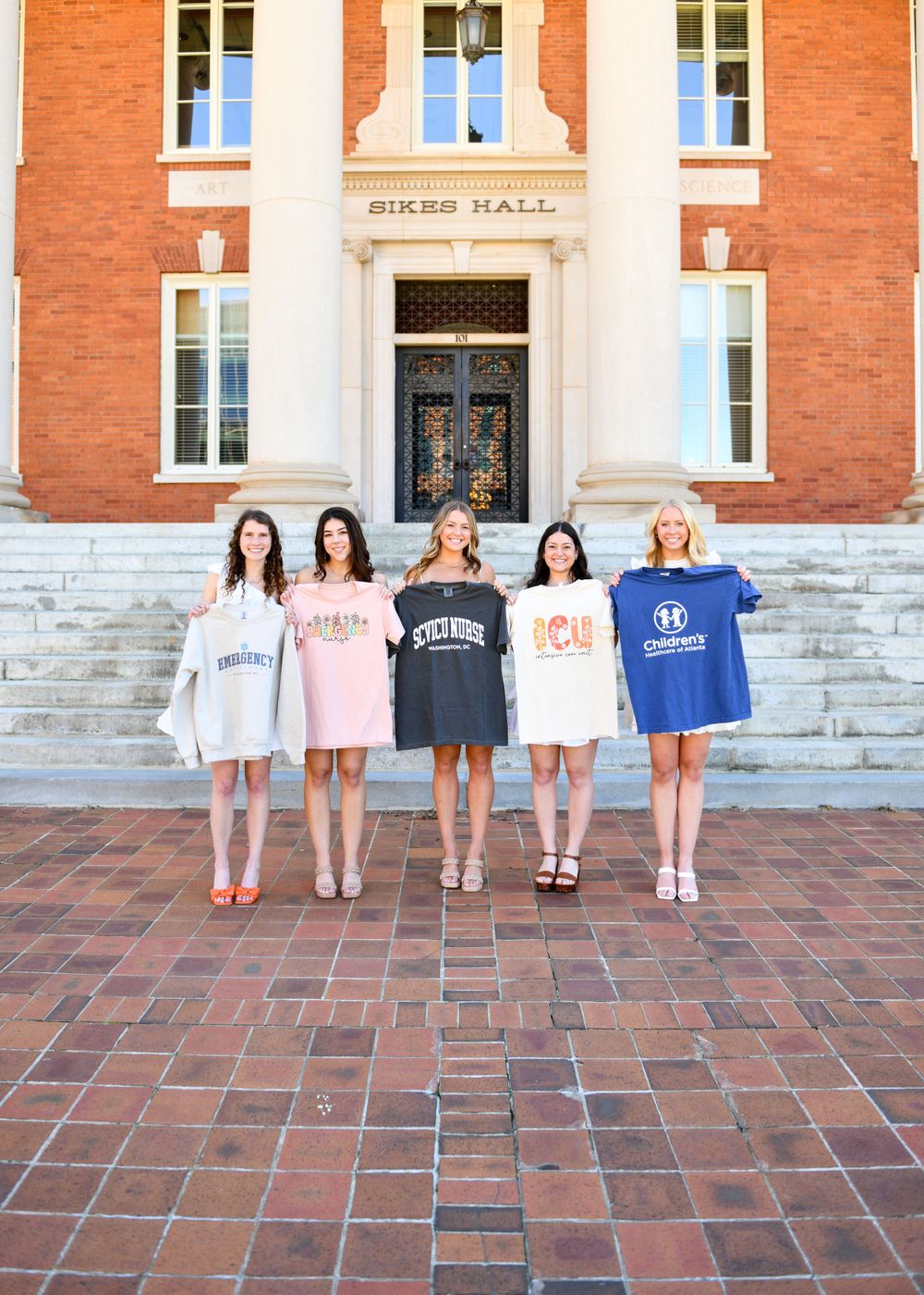 A group wearing college sweatshirts poses on brick steps in front of a historic building entrance.