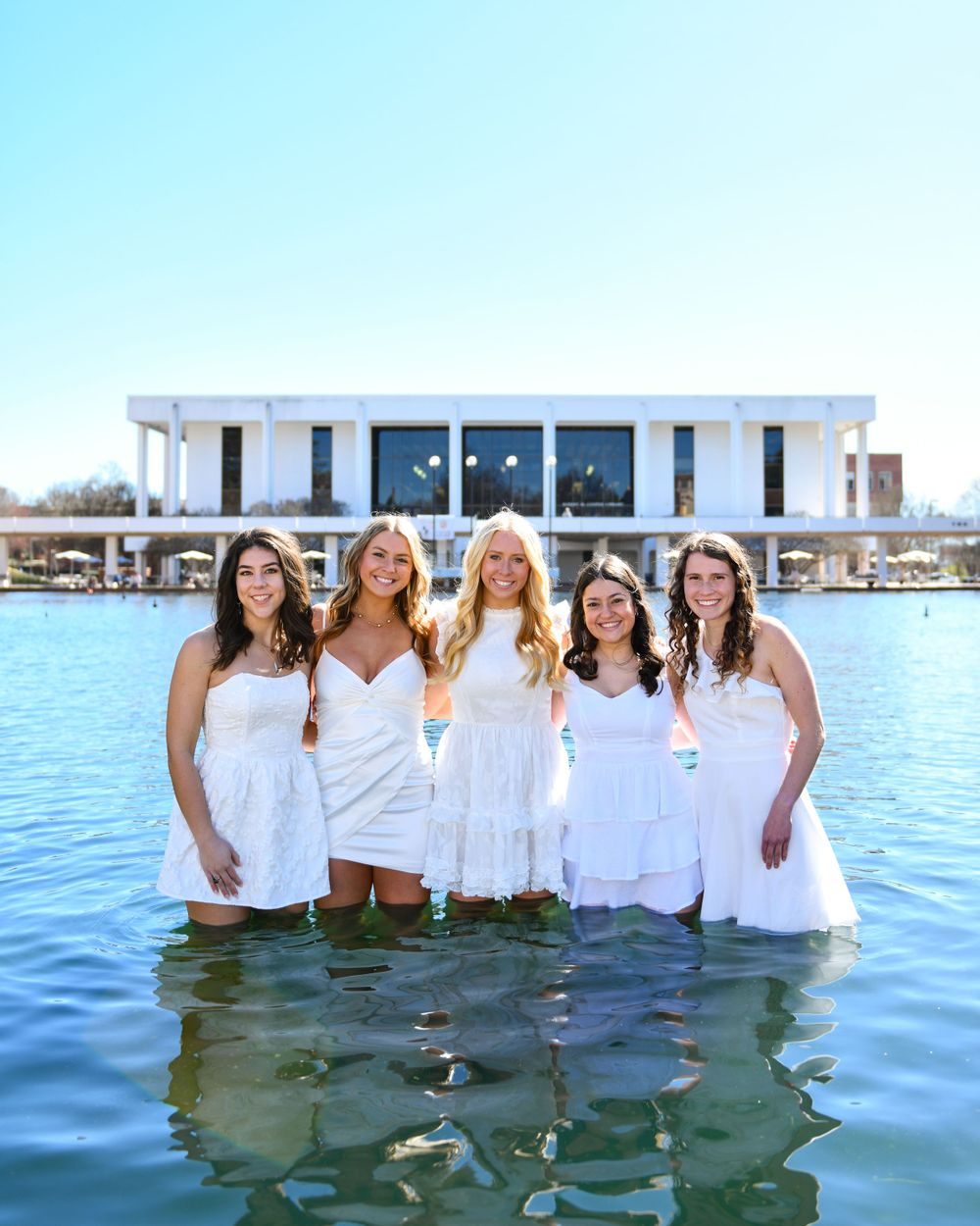 A group in white dresses stands in reflection pool water with a modern building in background.