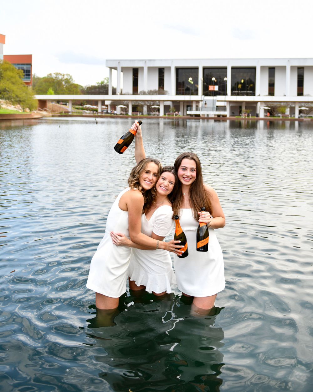 Friends celebrate in white dresses while splashing champagne in a reflective pool.