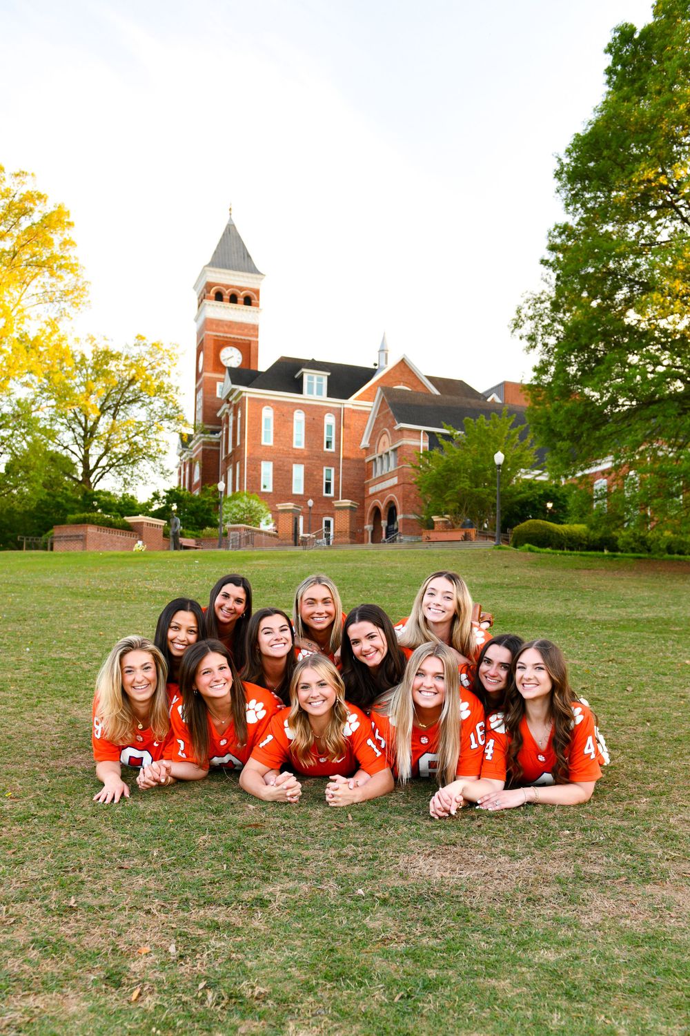 Students in orange shirts lie on grass in front of a classic brick university building with clock tower.