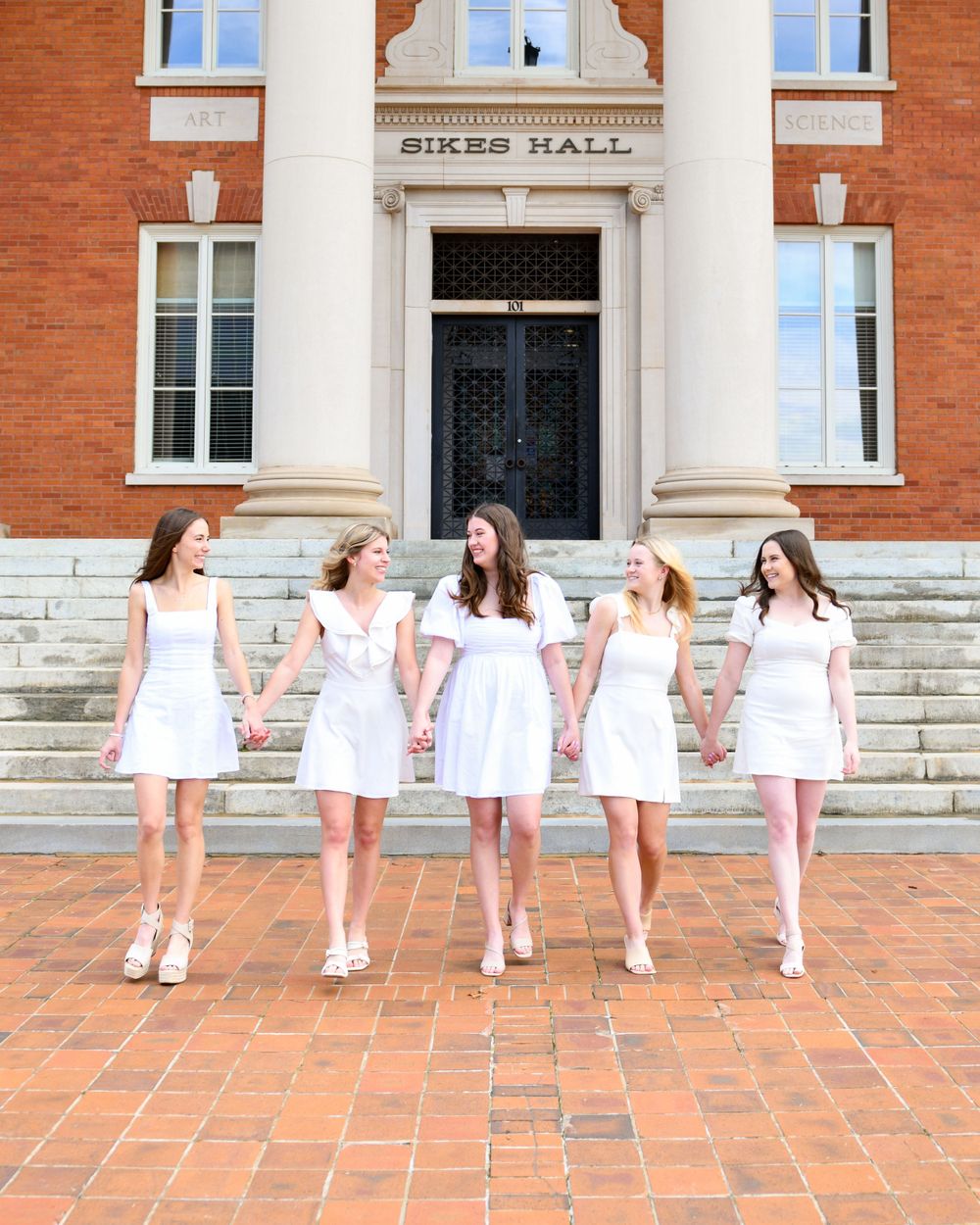 Five people in white dresses pose on brick steps in front of Sikes Hall at Clemson University.