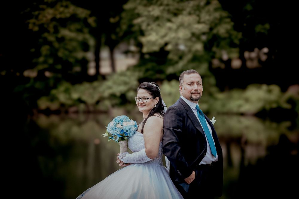 Bride and groom standing back-to-back outdoors near a pond, bride holding a blue bouquet in a white ball gown.
