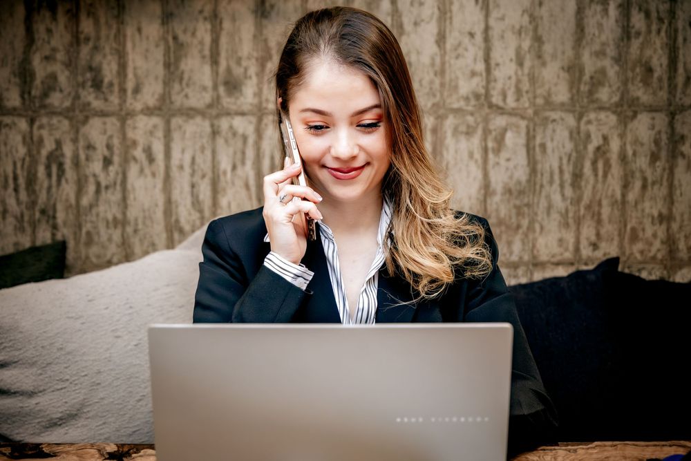 Young businesswoman smiling while talking on phone and working on laptop in professional setting.
