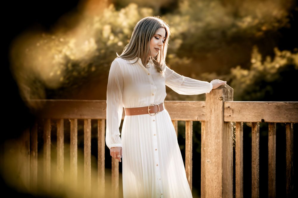Woman in flowing white dress and brown belt stands thoughtfully by wooden railing at golden sunset.