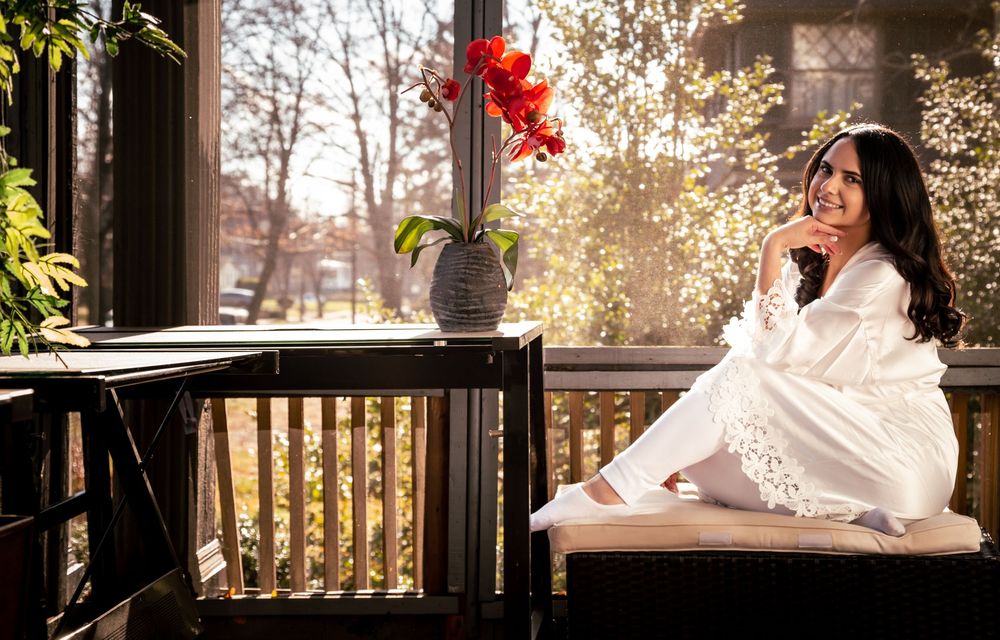 Woman in white outfit smiling on sunlit porch with red flower arrangement and autumn trees in background.