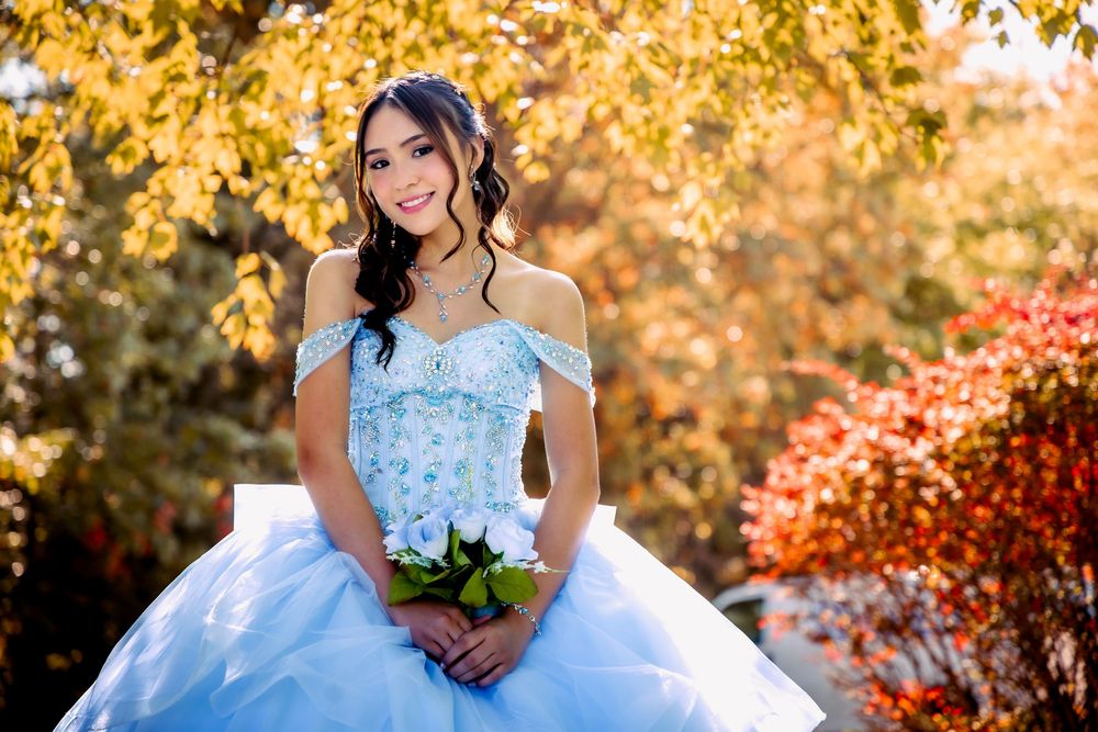 Young woman in a light blue off-shoulder quinceañera ball gown holding white roses outdoors in autumn.