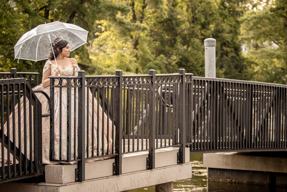 Bride in elegant gown holding white parasol umbrella on decorative bridge surrounded by lush green trees.