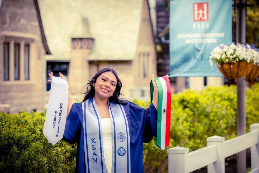 Kean University graduate in blue cap and gown holding flags outdoors near Hispanic serving institution sign.
