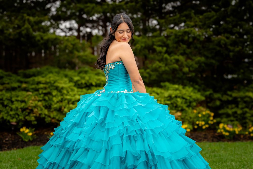 Young girl in a stunning teal ball gown with ruffled layers poses outdoors in a lush green garden setting.