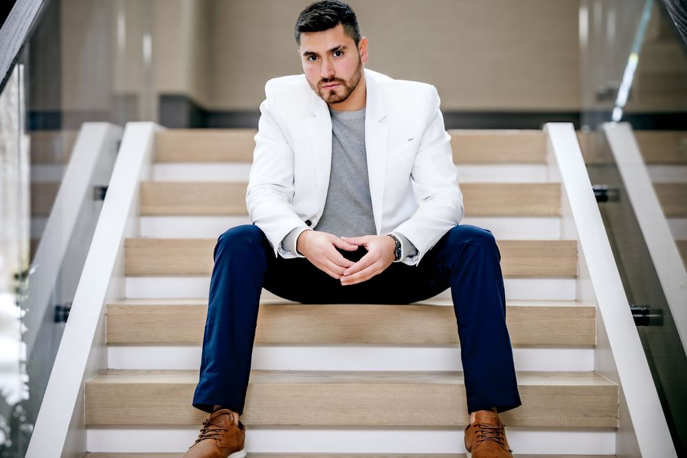Young man in white blazer and navy pants sitting on modern stairs with hands clasped, professional portrait photo.