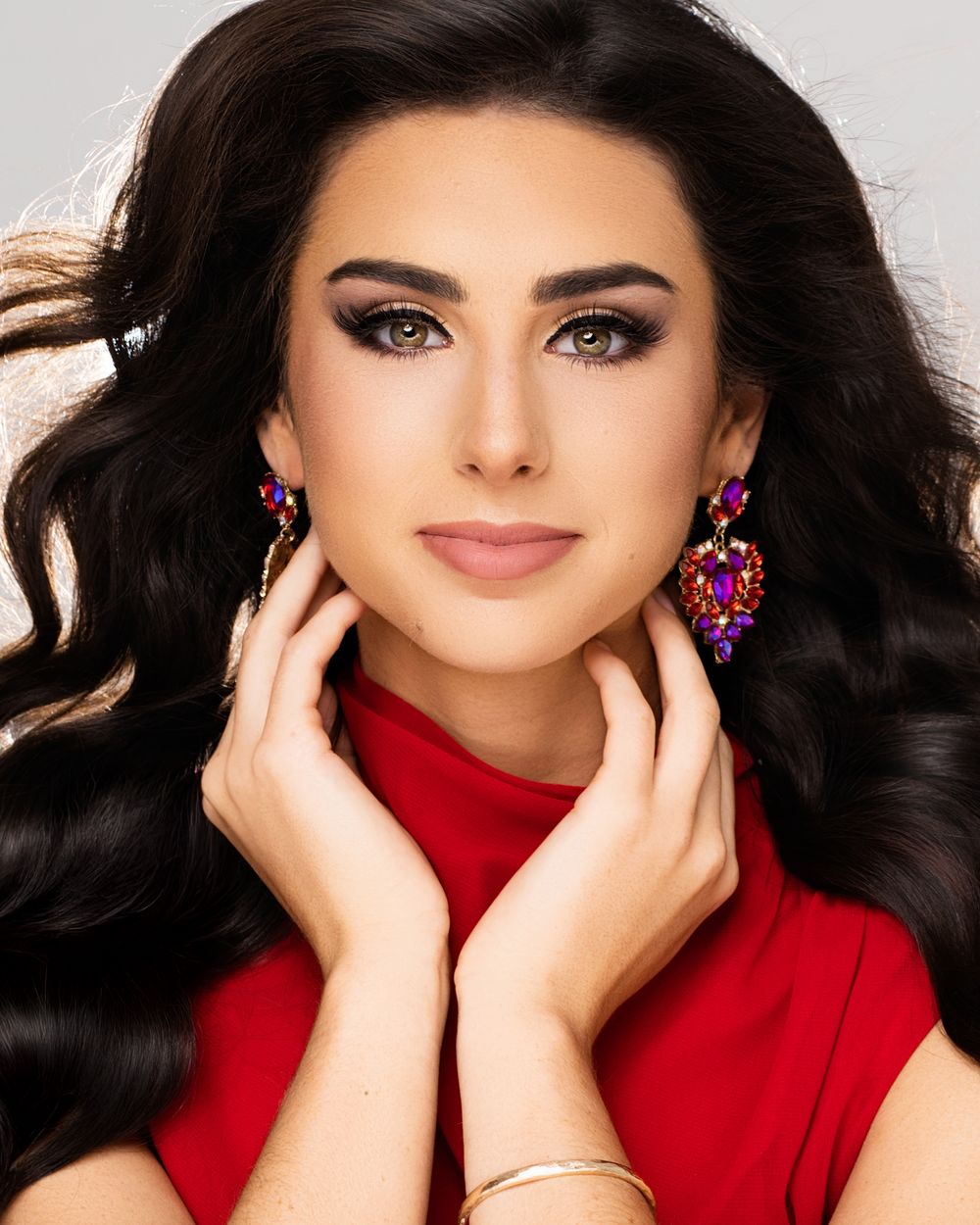 A glamour portrait of someone wearing a red top and statement pink earrings with dramatic eye makeup and flowing dark hair.