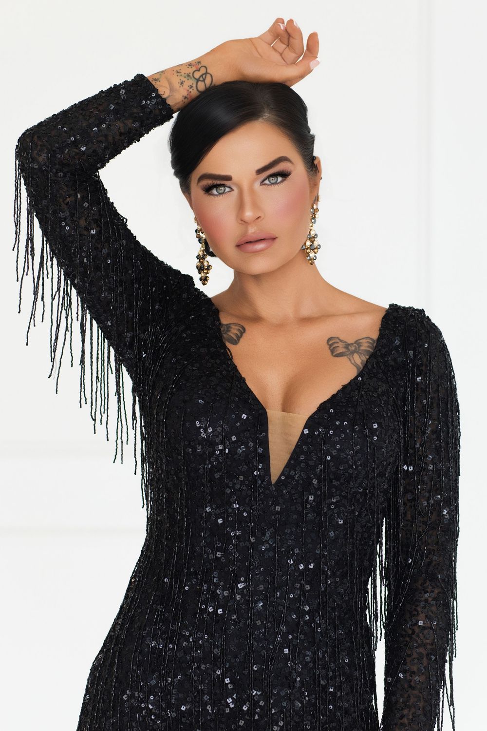 Woman in black sequined fringe dress with gold earrings and tattoos poses elegantly against white background.