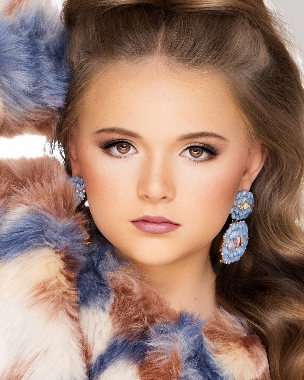 Close-up portrait featuring dramatic eye makeup, wavy brown hair, and sparkling blue crystal earrings against a soft background.